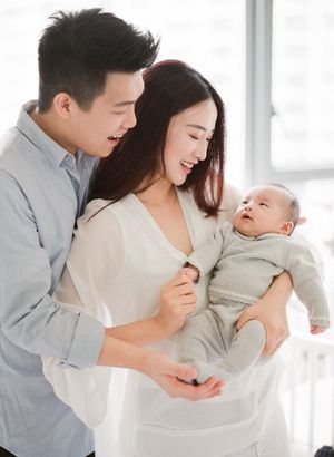 Happy young parents laughing together while holding their newborn baby by a bright window.