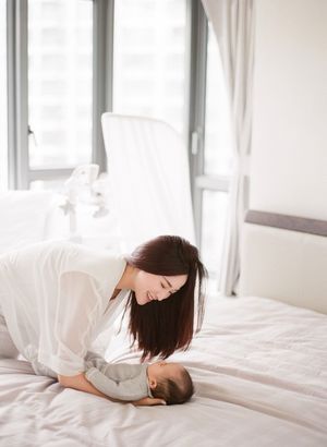 Mother in white shirt leaning over her sleeping newborn baby on a bright white bed.