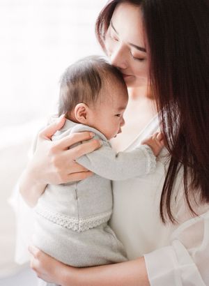 Tender moment of a mother gently kissing her newborn baby dressed in a gray knit outfit.