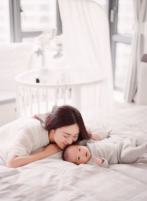 Mother nuzzling her smiling newborn baby lying on a white bed with a crib in background.