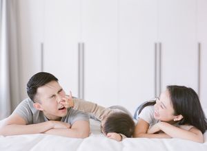 Young Asian family lying on a white bed laughing as baby touches fathers nose playfully.