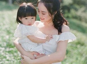 A young Asian mother in a white off-shoulder dress tenderly holds her smiling toddler outdoors.