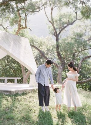 Happy Asian family holding hands with toddler in a sunlit park near a white structure.