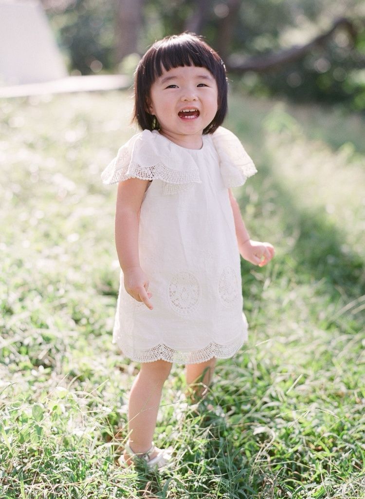 Cheerful Asian toddler girl in a white lace dress laughing and running on green grass.