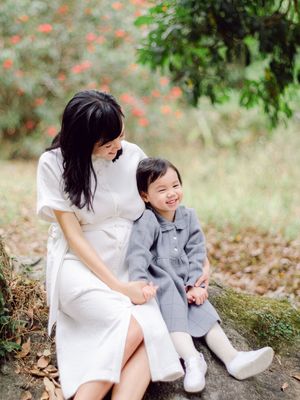 A woman and young girl in matching white and gray outfits sit together outdoors among green trees and red flowers.