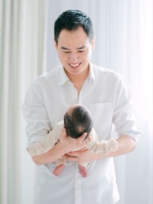 Smiling Asian father in a white shirt gently cradling his newborn baby near a bright window.