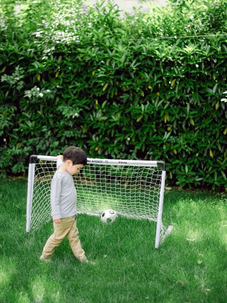Young boy playing soccer with a small goal net in a lush green backyard.