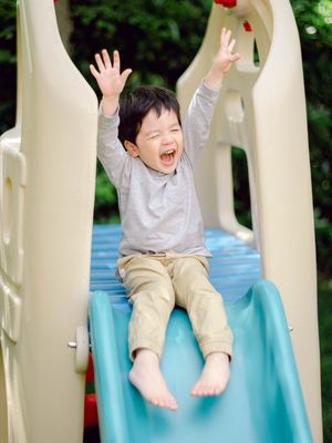 Joyful toddler boy with arms raised sliding down a playground slide.
