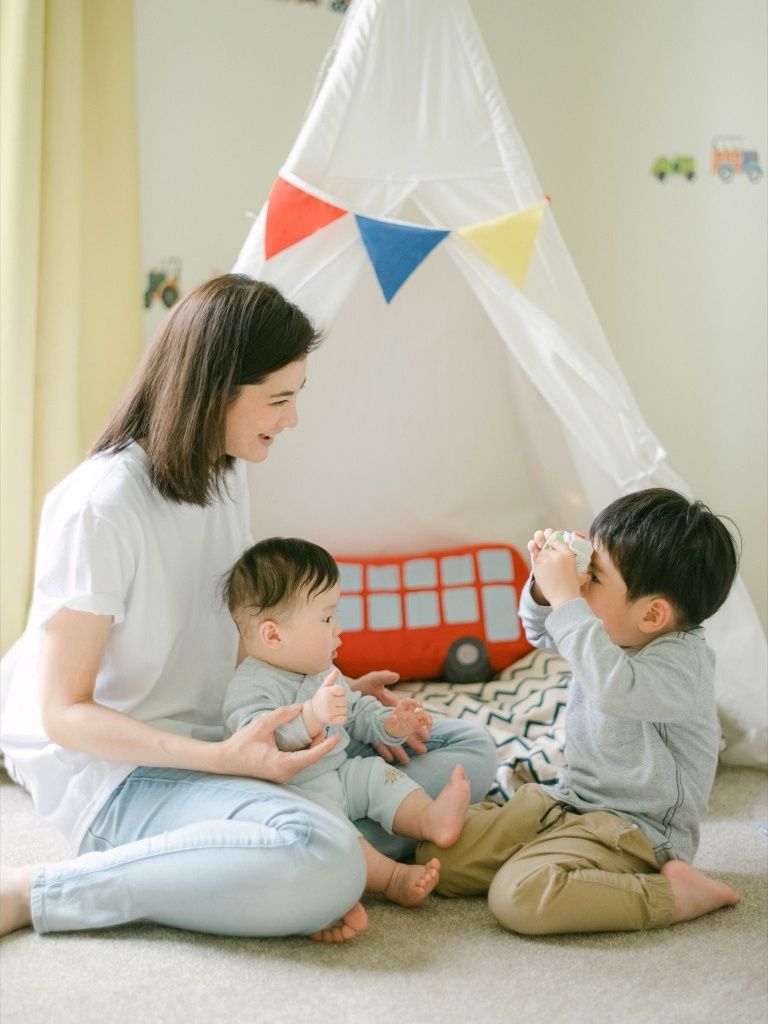 Mother sitting on floor playing with two young children near a teepee tent.