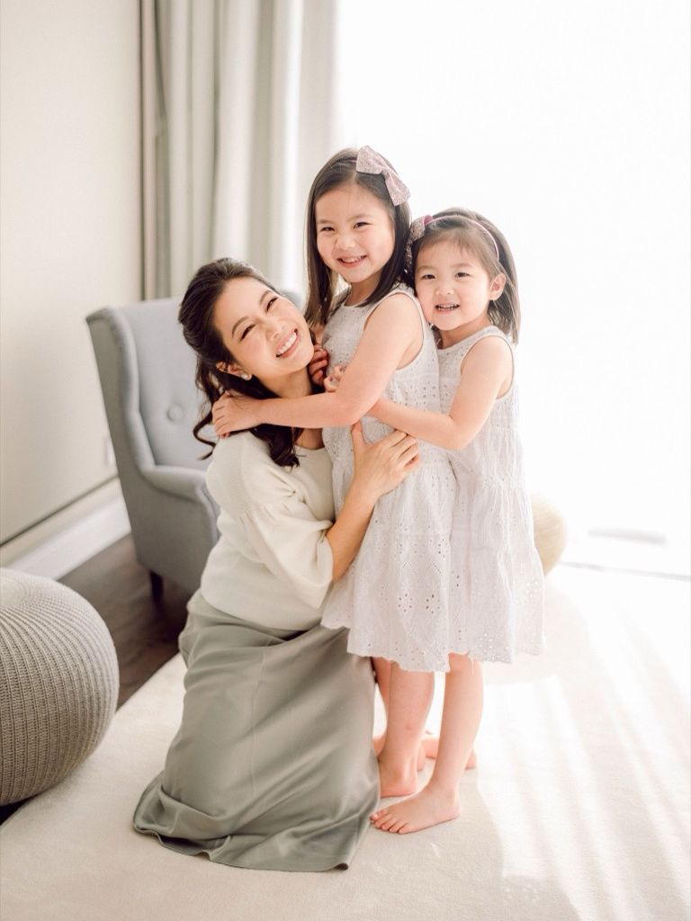 Happy mother kneeling and hugging two smiling young daughters in white dresses.