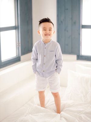Smiling young boy standing on a bed wearing a light blue button-up shirt.