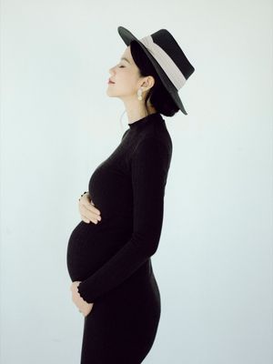 Elegant pregnant woman in black dress and hat cradling her baby bump.