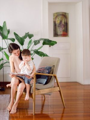 Mother sitting in a wooden chair reading a book with her toddler on her lap.