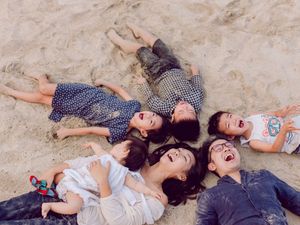 A joyful family of five lying together on sandy beach, laughing and looking up at the sky.