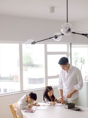 A father helps two young children with homework at a bright dining table in a modern home.