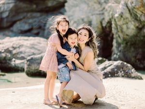 A smiling mother crouches on a rocky beach hugging her two laughing young children.