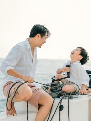 A father and young son share a laugh together while sitting on a sailboat on the ocean.