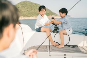 Two young boys playfully interact on the deck of a sailboat with scenic ocean views behind them.