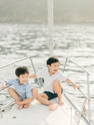 Two young boys sitting on the bow of a sailboat, smiling and laughing with sparkling water behind them.