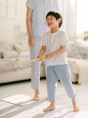 A smiling Asian boy plays with a toy bat indoors on a sunny day while an adult stands nearby.