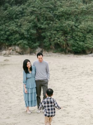 A couple in casual attire stands on a sandy beach as a young child runs toward them with trees behind.