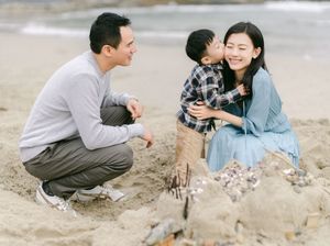 A family of three builds a sandcastle on the beach, with a boy kissing his mother on the cheek.