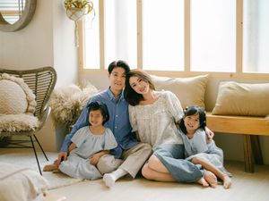A happy Asian family of four poses together on a cozy living room floor in soft blue and white outfits.