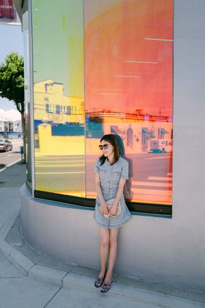 A young woman in a denim dress and sunglasses leans against a colorful rainbow glass storefront on a city street.