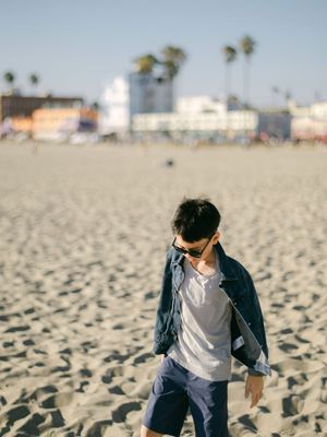 A young man in a denim jacket and sunglasses walks along a sandy beach with buildings in the background.
