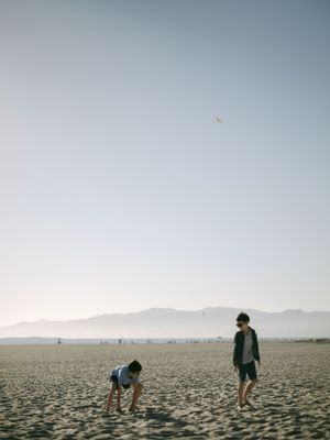 Two children play on a vast sandy beach under a hazy sky with mountains visible in the distance.