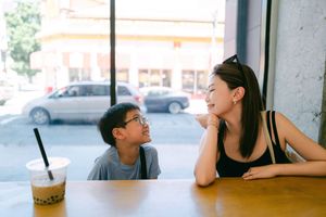 A woman and young boy share a warm conversation over bubble tea at a sunny cafe window seat.