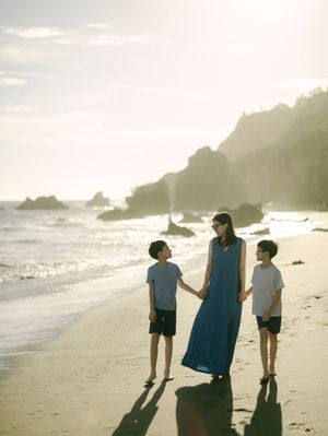 Woman in blue dress holds hands with two boys on a sunny beach with rocky cliffs.