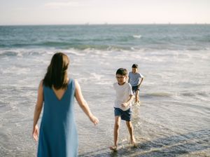 Woman in blue dress watches two boys playing in ocean waves at the beach.