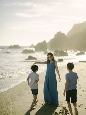 Mother in blue maxi dress walks with two young boys along a sunlit beach.