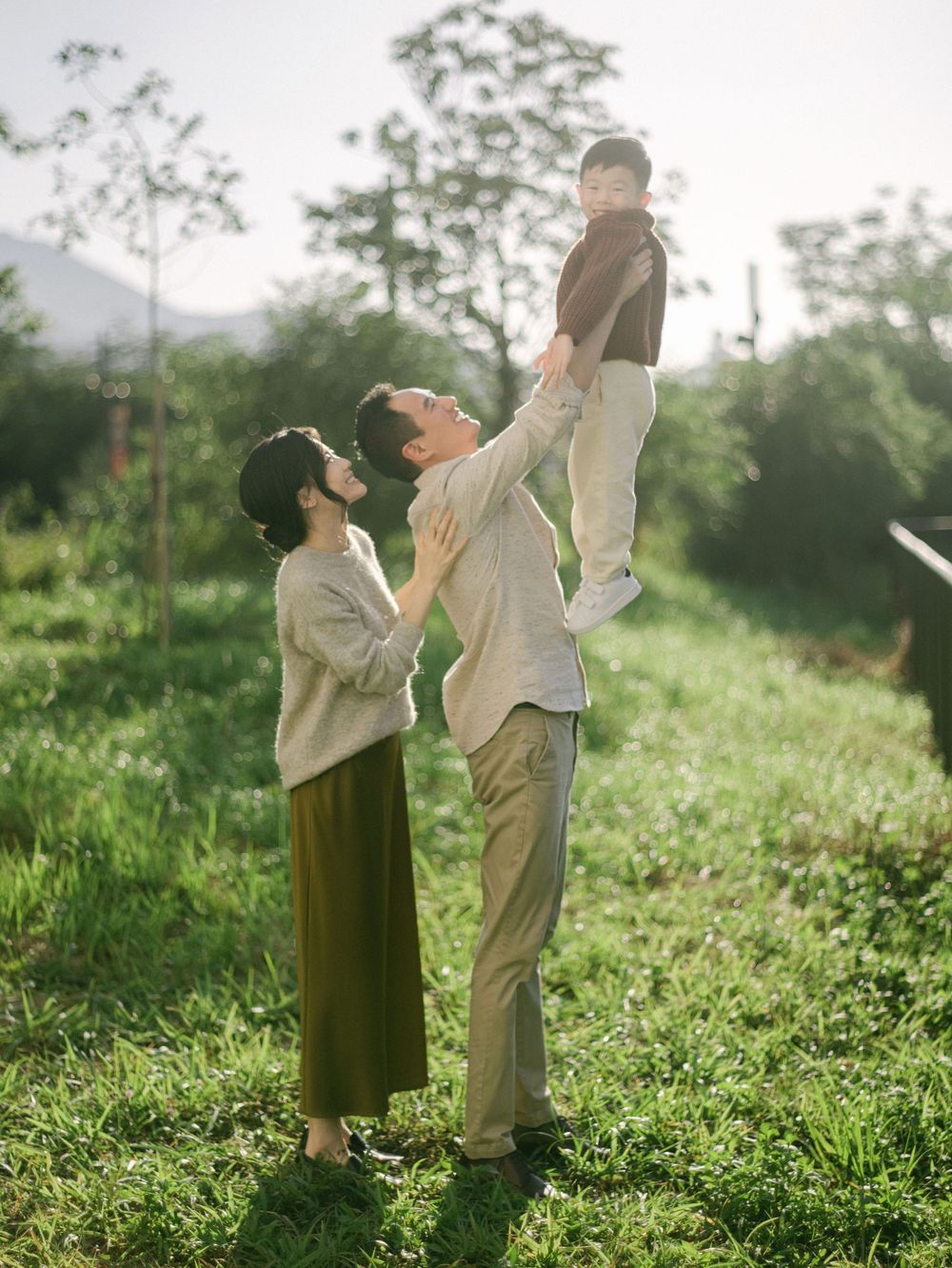 Father lifts smiling baby overhead while mother watches in a green sunlit field.