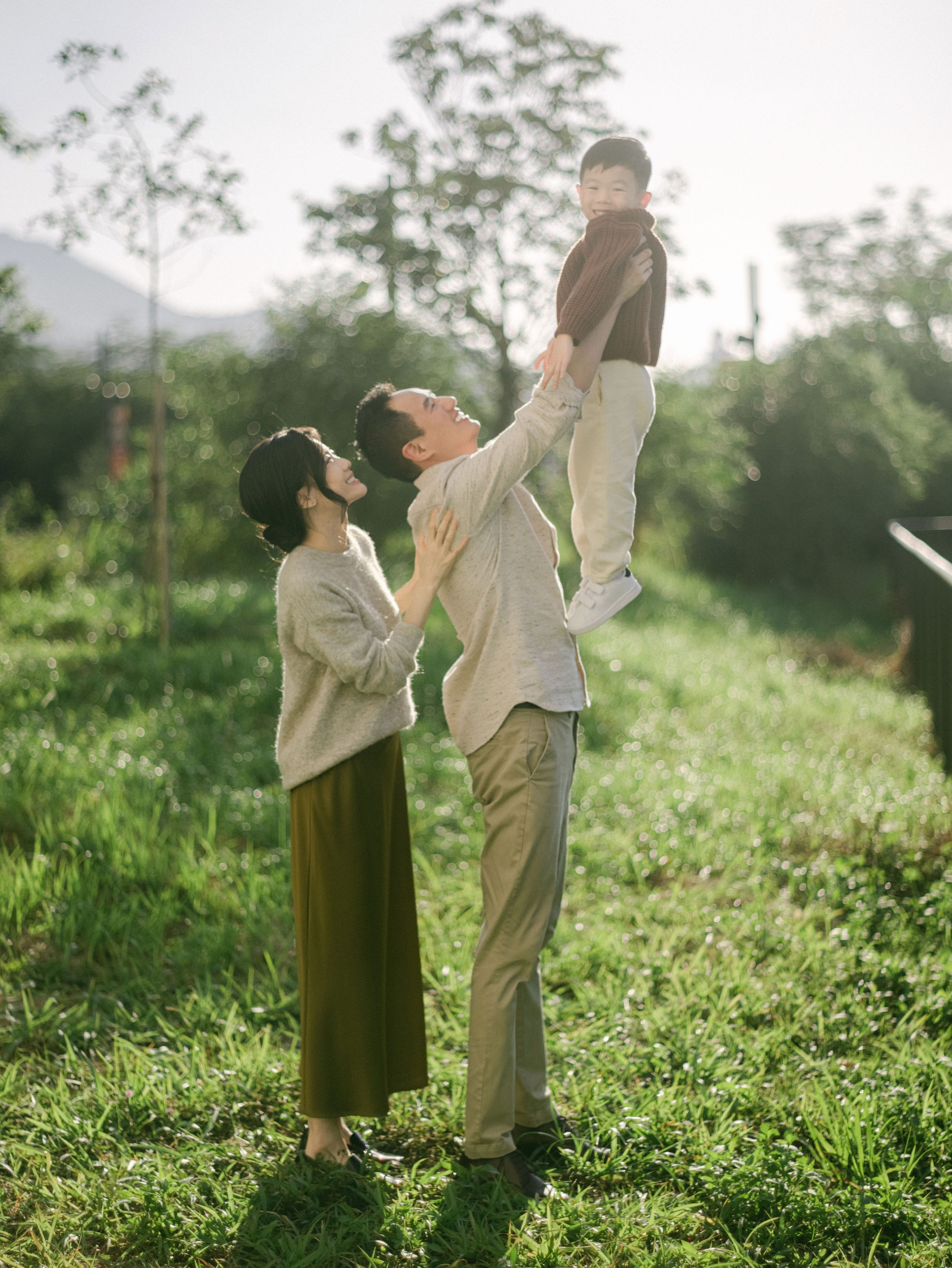 Father lifts smiling baby overhead while mother watches in a green sunlit field.