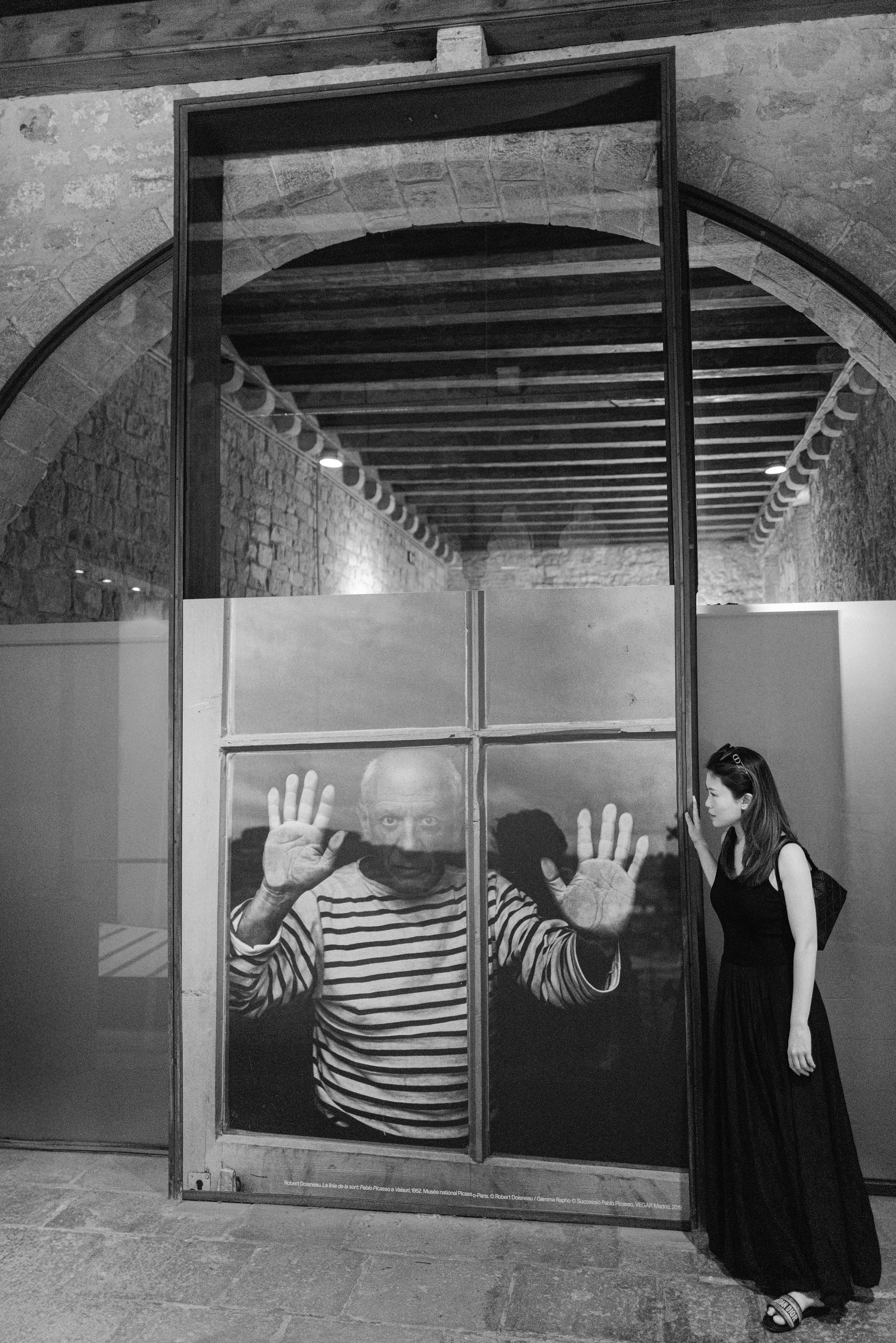 Woman in black dress looks at elderly man pressing hands on glass in B&W.
