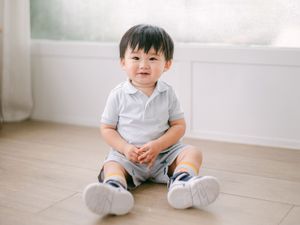 Adorable Asian toddler boy sitting on wooden floor smiling in a bright room.