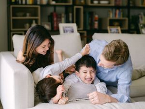 Happy family playing together on a white sofa, parents and two children laughing in a cozy living room.