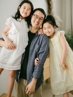 Smiling Asian mother posing with two young daughters in white and yellow dresses in a bright room.