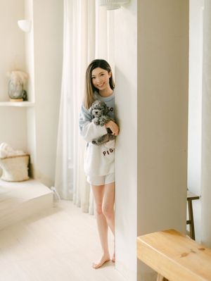 Young woman smiling and holding a grey toy poodle while leaning against a white wall indoors.