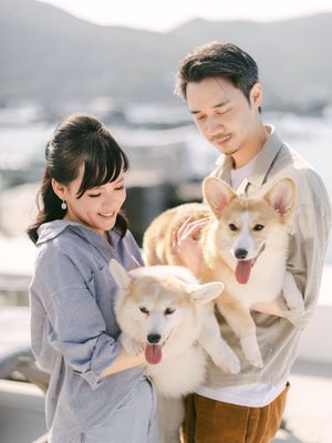 Smiling couple holding two fluffy Corgi puppies outdoors with a scenic background.