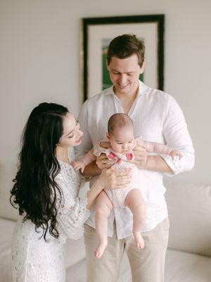 Happy young parents in white outfits smiling and holding their newborn baby indoors.