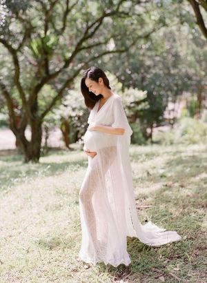 A pregnant woman in a flowing white gown stands gracefully in a sunlit outdoor garden.