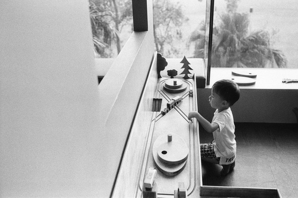 Black and white photo of a young child playing with a wooden train set by a large window.