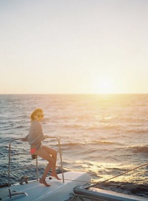 Woman standing on sailboat bow watching golden sunset over ocean waves.