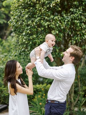 Happy couple lifting smiling baby in lush green garden setting.
