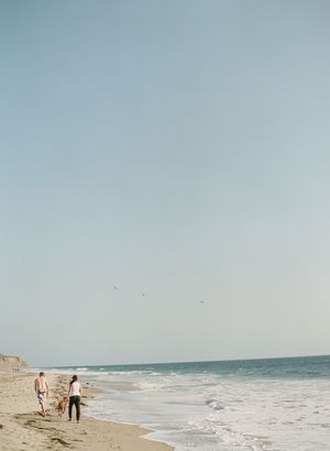 Couple walking with dog along sandy beach with clear blue sky.