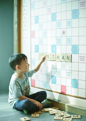 A young boy spelling his name ETHAN on a giant Scrabble board with wooden letter tiles.