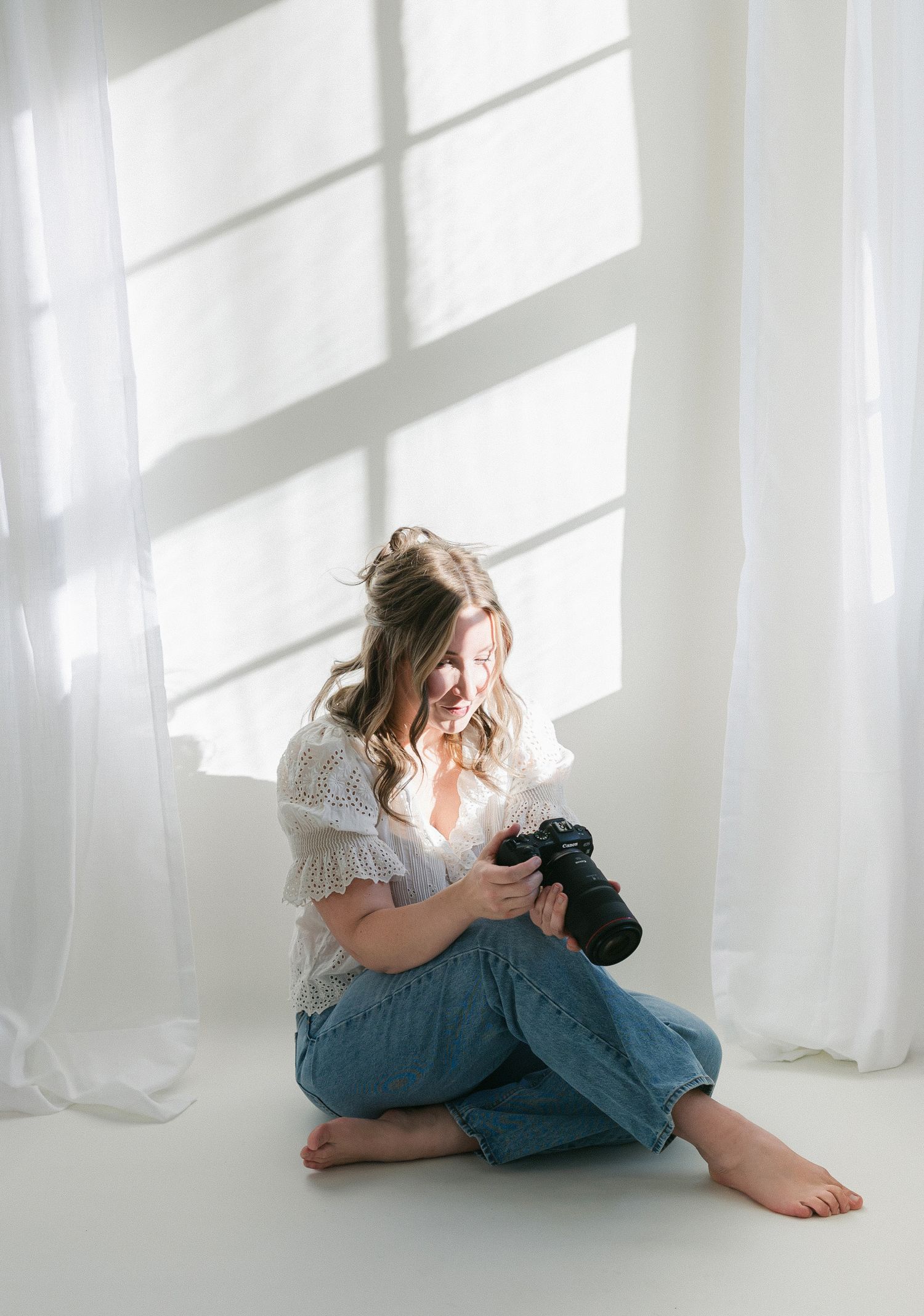 A barefoot woman sits cross-legged on a white studio floor, smiling at her camera, wearing a white top and blue jeans.